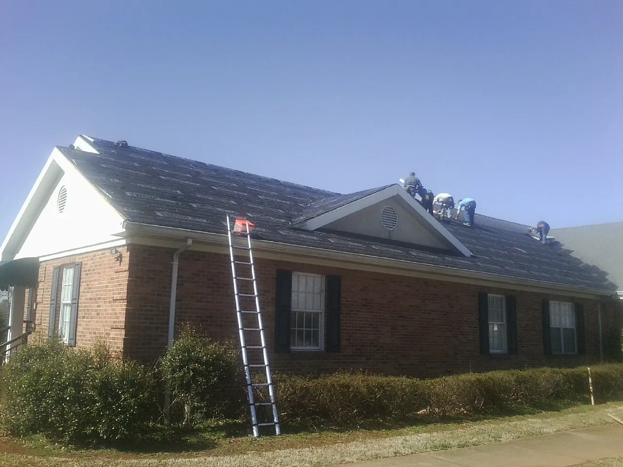 Crew tearing shingles off a brick ranch roof with a ladder set — Emergency Roof Repair work by our crew in Doucette