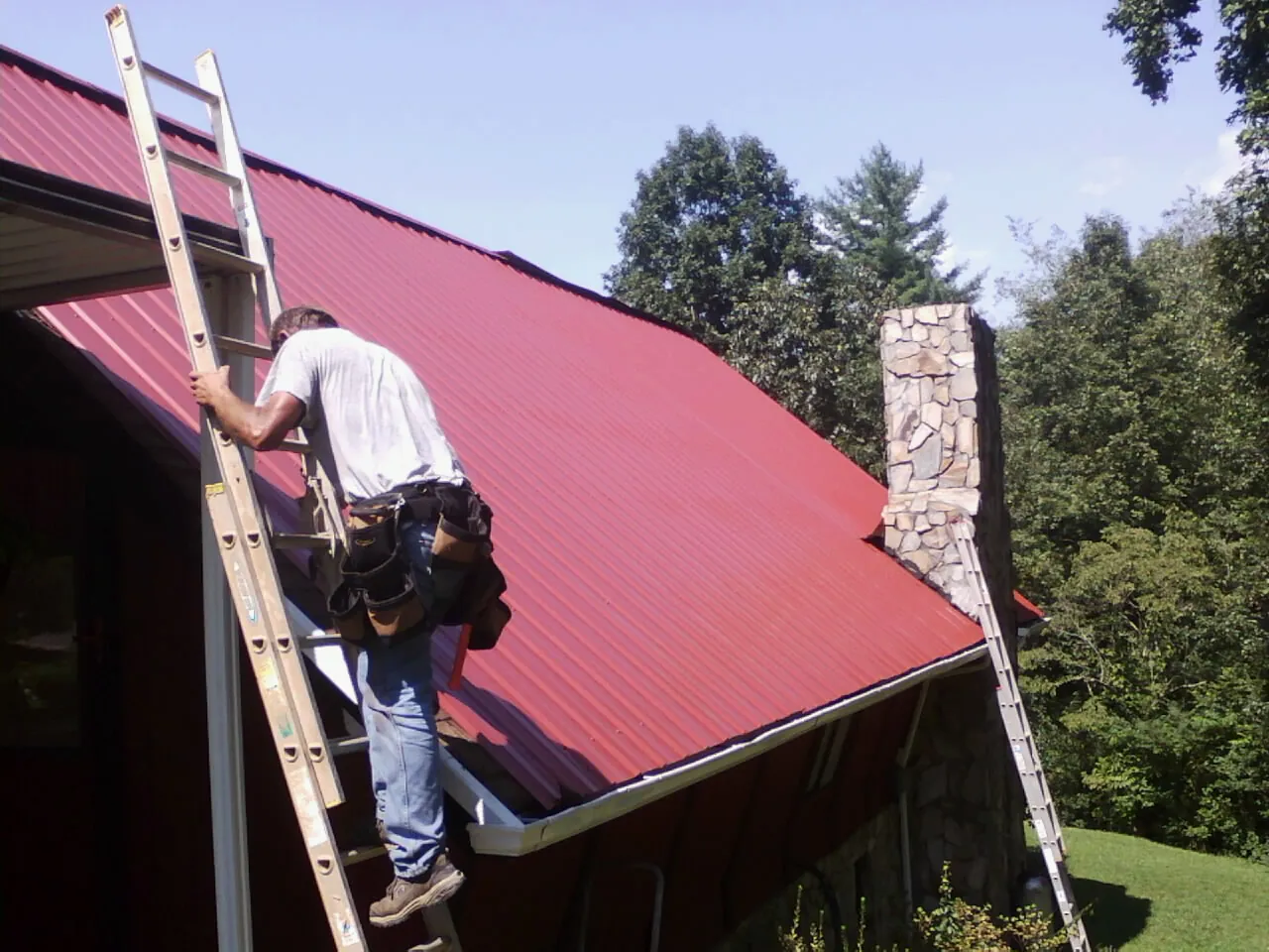 Roofer on a ladder beside a red metal roof and stone chimney — Soffit & Fascia Repair by Roofing Brothers in Doucette